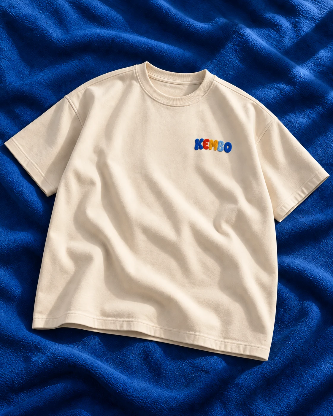 Front concept of an ecru oversized tee with a small colourful KEMBO logo on the left chest, on cobalt blue fabric